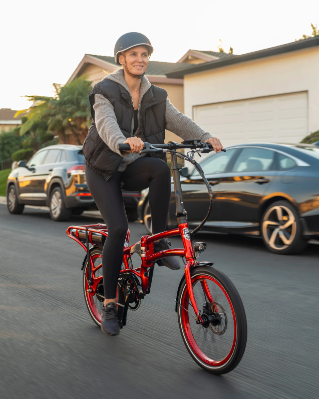 Person riding a red Pedego Latch on a residential street with cars and houses in the background