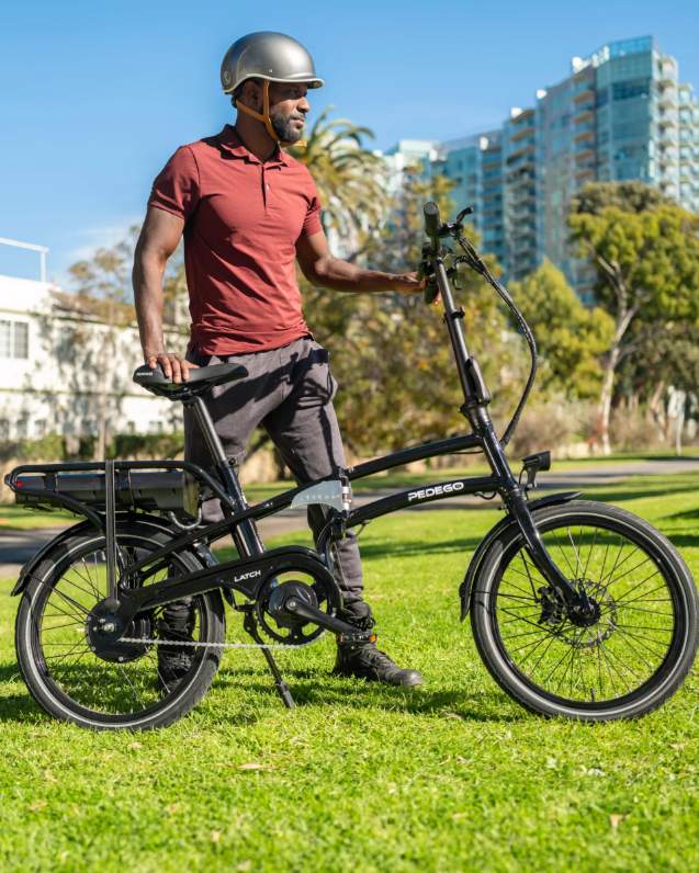 Man with a helmet standing next to a black Pedego Latch V2 e-bike in an urban park setting.